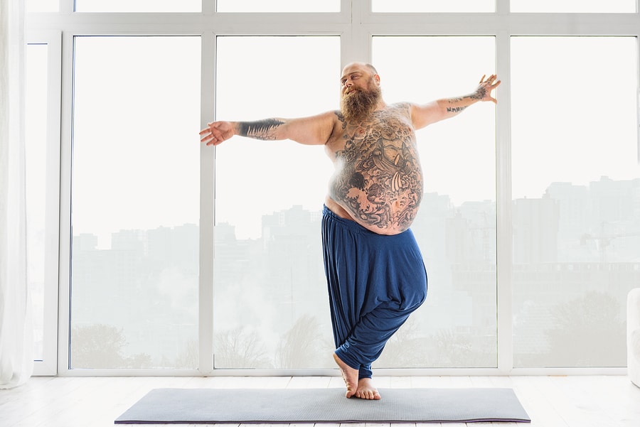 Graceful fat man is doing yoga with pleasure. He is stretching arms sideways and relaxing. Man is standing on mat and smiling
