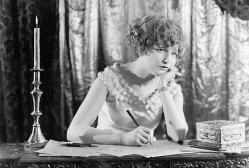 A woman sits and thinks at a desk with pen in hand as if she is deciding whether to outsource content writing.